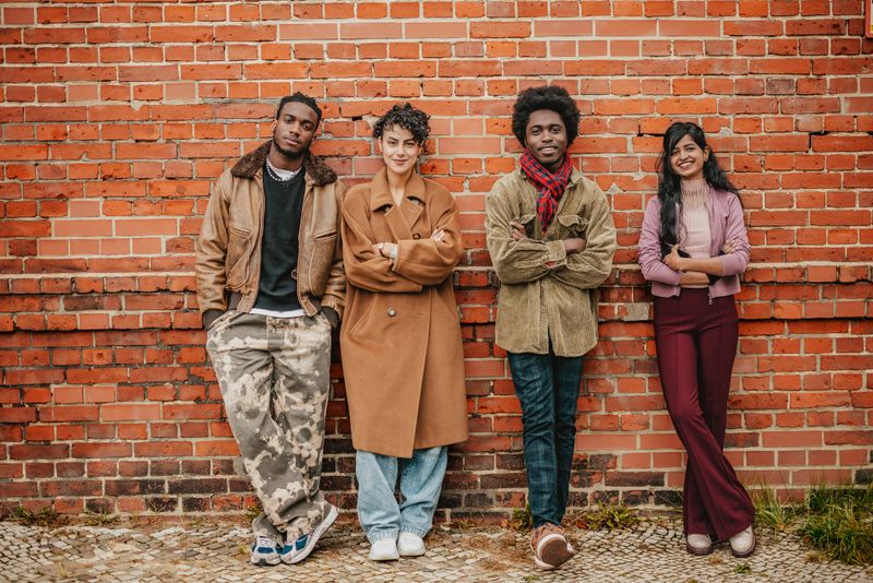 Four young adults stand against a brick wall in Berlin, each displaying their unique style and joyful demeanor. They are enjoying their time outdoors in the city while sharing smiles and laughter.