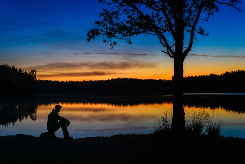 A man observes the calm water of a lake as the sun sets, creating a beautiful scene of color and tranquility. The silhouette contrasts against the twilight sky near Gothenburg.