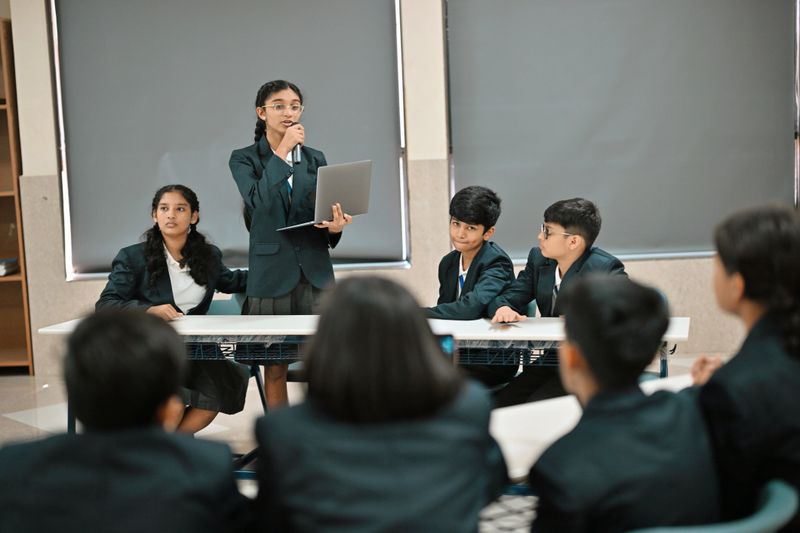 Students in formal uniforms sit in teams while the girl participant stands with a Laptop and a microphone to present her arguments, as the opposing team listens and prepares their rebuttal.
