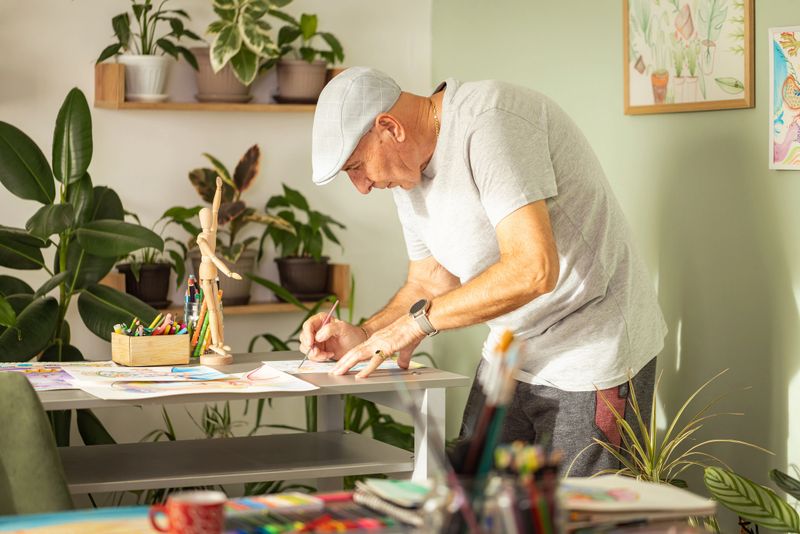 An engaged senior artist, surrounded by lush houseplants, meticulously works on an illustration at his bright home desk, embodying a serene and productive retirement.