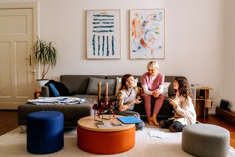 Three female friends relax in a living room, with one wearing medical scrubs, using a smartphone. Captures friendship, casual indoor lifestyle, and modern personal interaction.