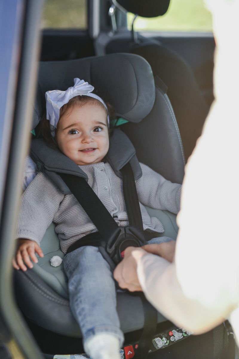 A cheerful toddler sits securely in a rear-facing car seat, wearing a soft gray sweater and a white bow headband. The scene conveys warmth, safety, and loving family moments during a car trip.