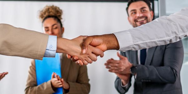 Two people shaking hands while others applaud in a business setting.