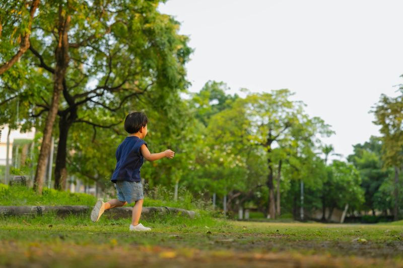happy Asian toddler boy runs playfully through a green garden, laughing with pure joy as he explores trees and nature. His energetic movement reflects fun, curiosity, and healthy childhood exercise in a bright outdoor park setting filled with life and wonder