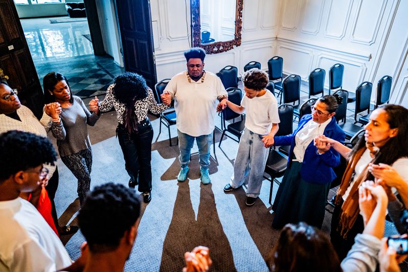 A dynamic group prayer scene captures intense spiritual moment, with participants holding hands and expressing deep emotional connection in a beautifully decorated interior with vibrant blue, orange, and white architectural elements and elegant chandeliers.