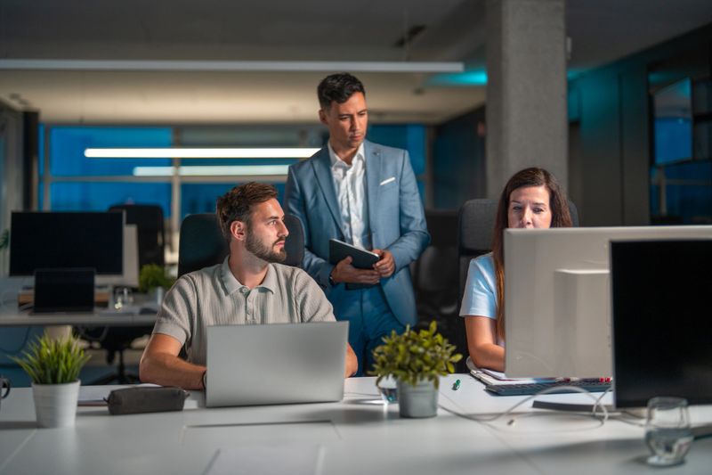 Mid-adult Japanese manager attentively guiding a group of diverse business executives working late in a modern office setting as they focus on meeting an approaching deadline, fostering collaboration and efficiency in the workplace.