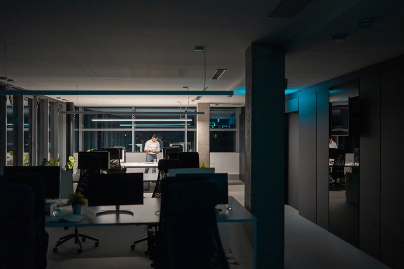 Focused mid-adult Asian man working late at night in a modern open-plan office, contemplating at his desk. The quiet atmosphere highlights his dedication and concentration as he navigates through tasks under the office lights.
