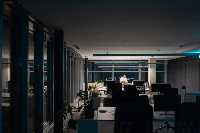 Focused mid-adult Asian man working late in an open-plan office while contemplating tasks at his desk, surrounded by sleek modern design and soft lighting.