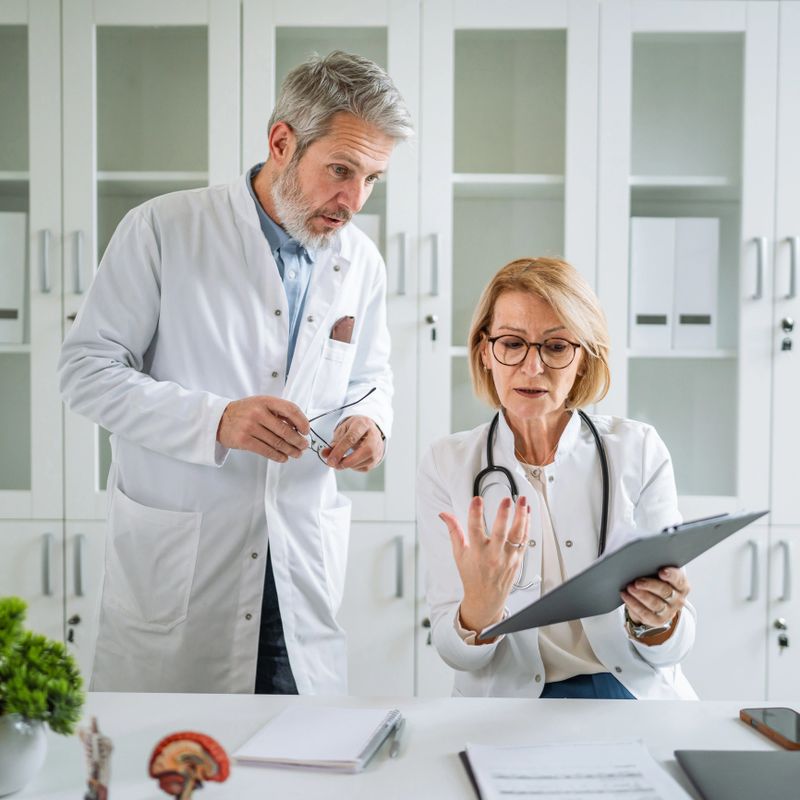 Medical professionals, a mature man and woman, smiling and collaborating while reviewing patient records and discussing healthcare information with a clipboard in a bright office environment