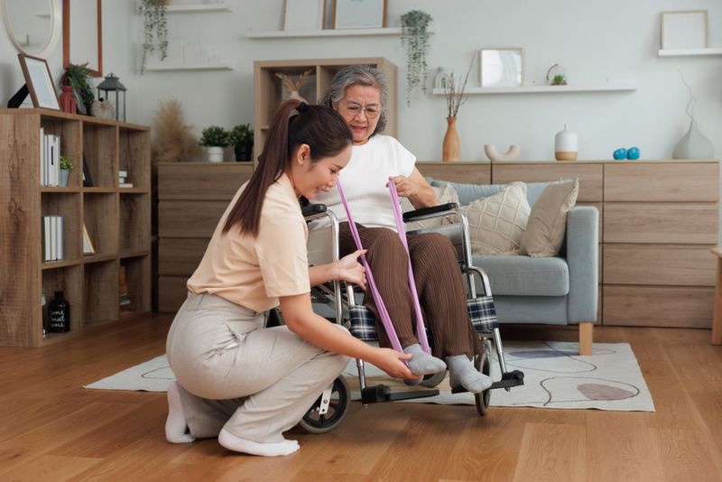 A caregiver assists an elderly woman in a wheelchair with a leg stretch using a resistance band in a bright, cozy living room. This scene represents physical therapy, mobility support, rehabilitation, senior fitness, home healthcare, and compassionate caregiving. Ideal for concepts of aging well, therapist support, assisted exercise, occupational therapy, and home-based elder care.