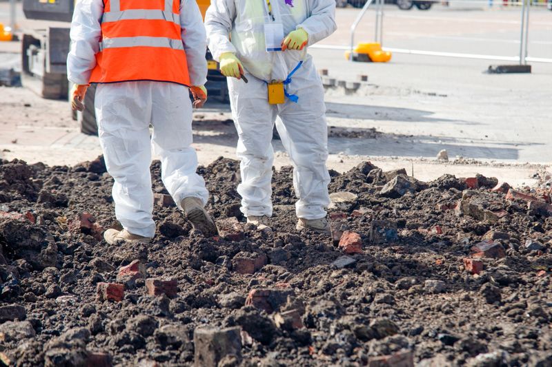 Two construction workers in safety gear inspect a construction site covered with broken soil and bricks. It is a clear day, indicating ongoing urban development.