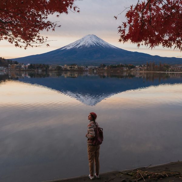 Person admiring Mount Fuji across a calm lake framed by autumn leaves.