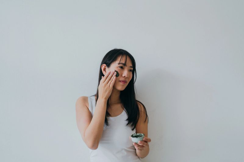 Photo of a young Asian woman applying homemade facial mask, she's just made at her home out of a natural, organic ingredients: clay and matcha