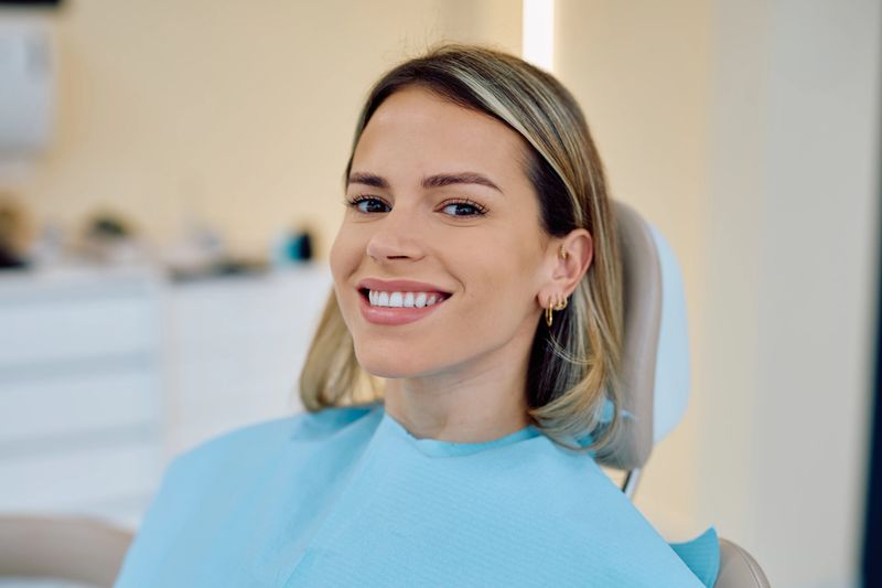 Smiling young woman sitting in a dental chair, wearing a protective bib. She is happy with her healthy teeth