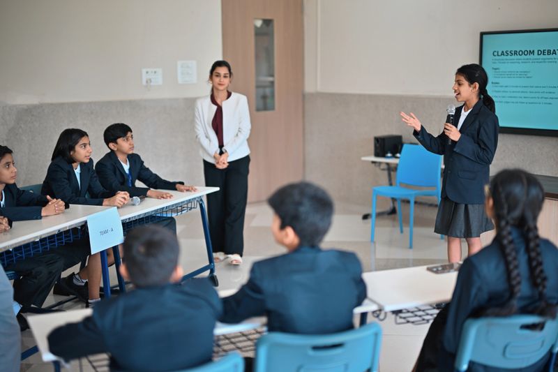 A confident middle school student moderates a classroom debate, speaking into a microphone as two uniformed teams listen attentively. The teacher stands nearby observing. Captures student leadership, public speaking, teamwork, and active learning in a modern school setting.
