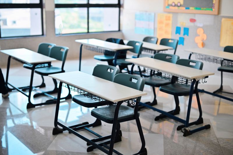 Rows of modern student desks and chairs sit neatly arranged in a bright classroom with large windows and wall posters. The tidy, unused space evokes back-to-school themes, learning environments, exam readiness, and education infrastructure. Ideal for illustrating schools, teaching, timetables, or classroom management concepts.