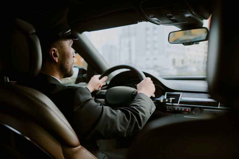 Caucasian middle aged man driving car in urban setting, holding steering wheel with both hands, wearing formal attire, focused on road ahead, city buildings visible through windshield