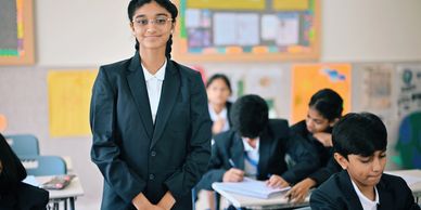 Confident schoolgirl standing in classroom with classmates studying behind her.