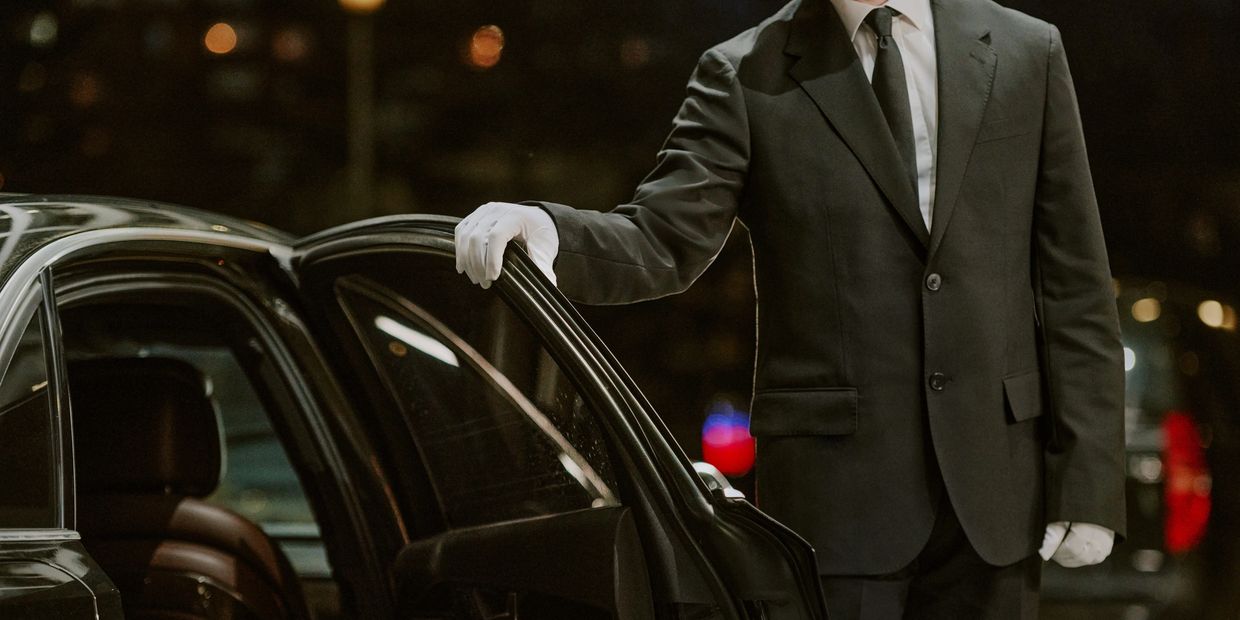 A chauffeur in a black suit and white gloves holding a car door open.