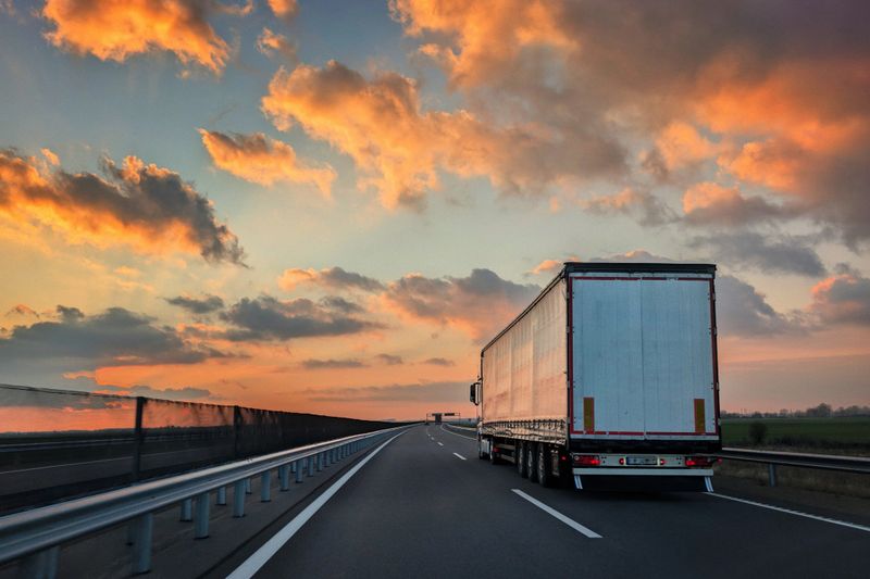 This image captures the rear view of a large, white commercial semi-truck and trailer driving away down a dark asphalt highway, with the truck centered slightly to the right of the frame. The scene is illuminated by the warm, dramatic light of a sunrise or sunset, featuring a sky filled with deep blue, orange, and golden clouds, and the roadside includes a metallic guardrail and a wire mesh fence extending into the distance. This photograph is commercially useful for themes related to transportation, freight logistics, long-haul shipping, road infrastructure, time-sensitive delivery, and the trucking industry.