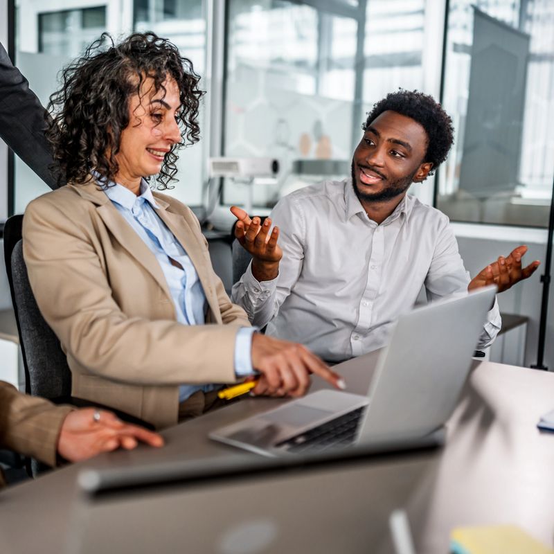 Diverse business professionals discussing work, using laptop, and sharing ideas in a modern corporate office environment