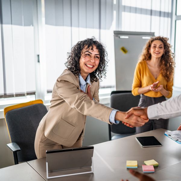 A smiling woman in a beige suit shaking hands in a meeting room.