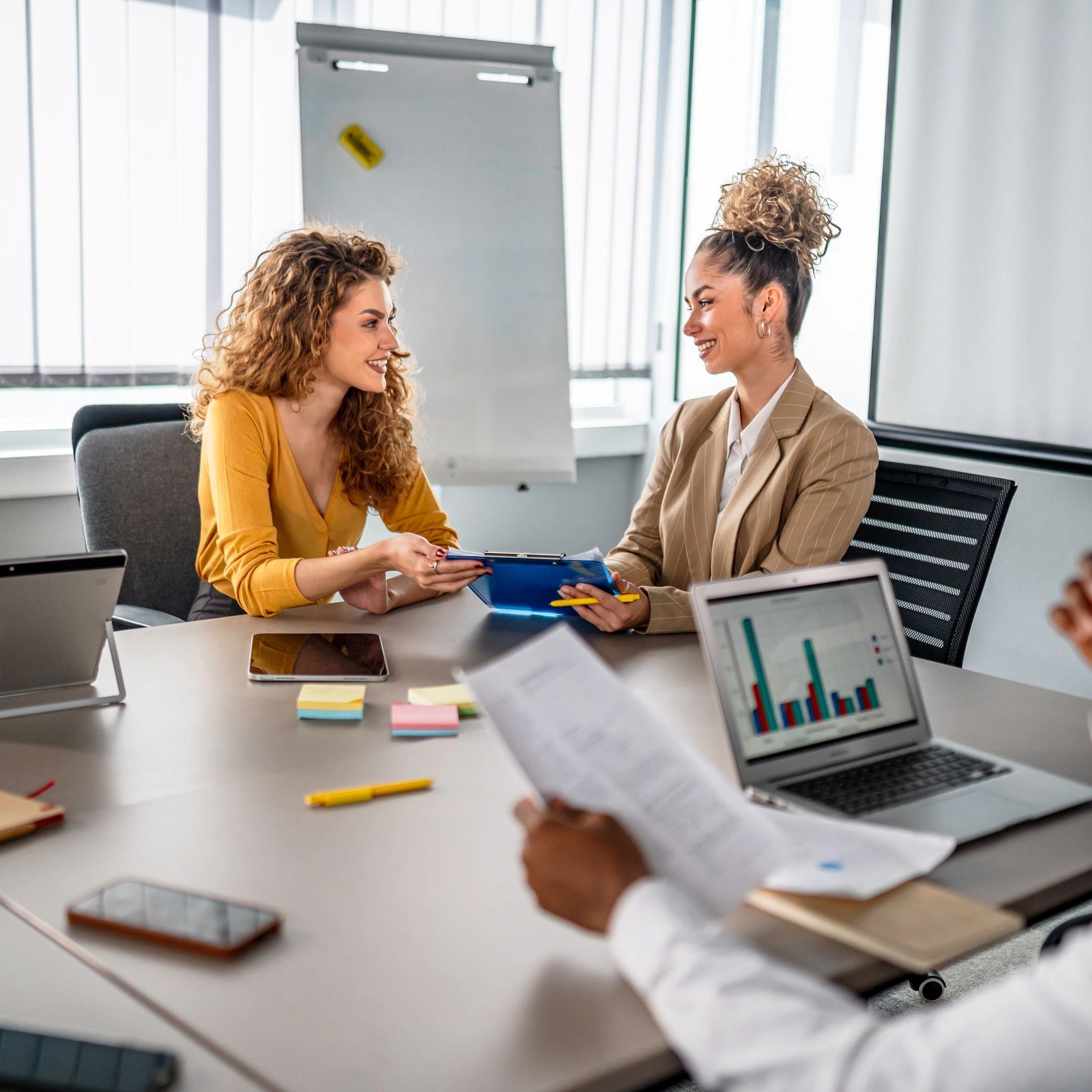 Two women smiling and sharing a document during a meeting in a bright office.