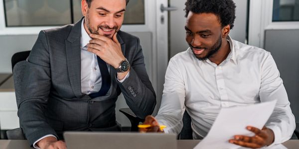 Two businessmen discussing work in an office with a laptop and documents.