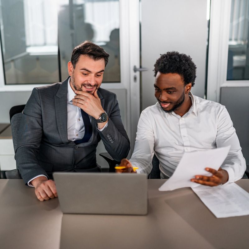 Two businessmen are collaborating on a laptop in an office, reviewing documents and discussing a project with smiles