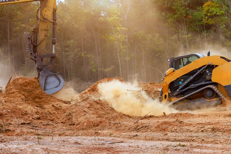 Two pieces of heavy machinery are busy clearing dirt debris at construction site surrounded by trees.