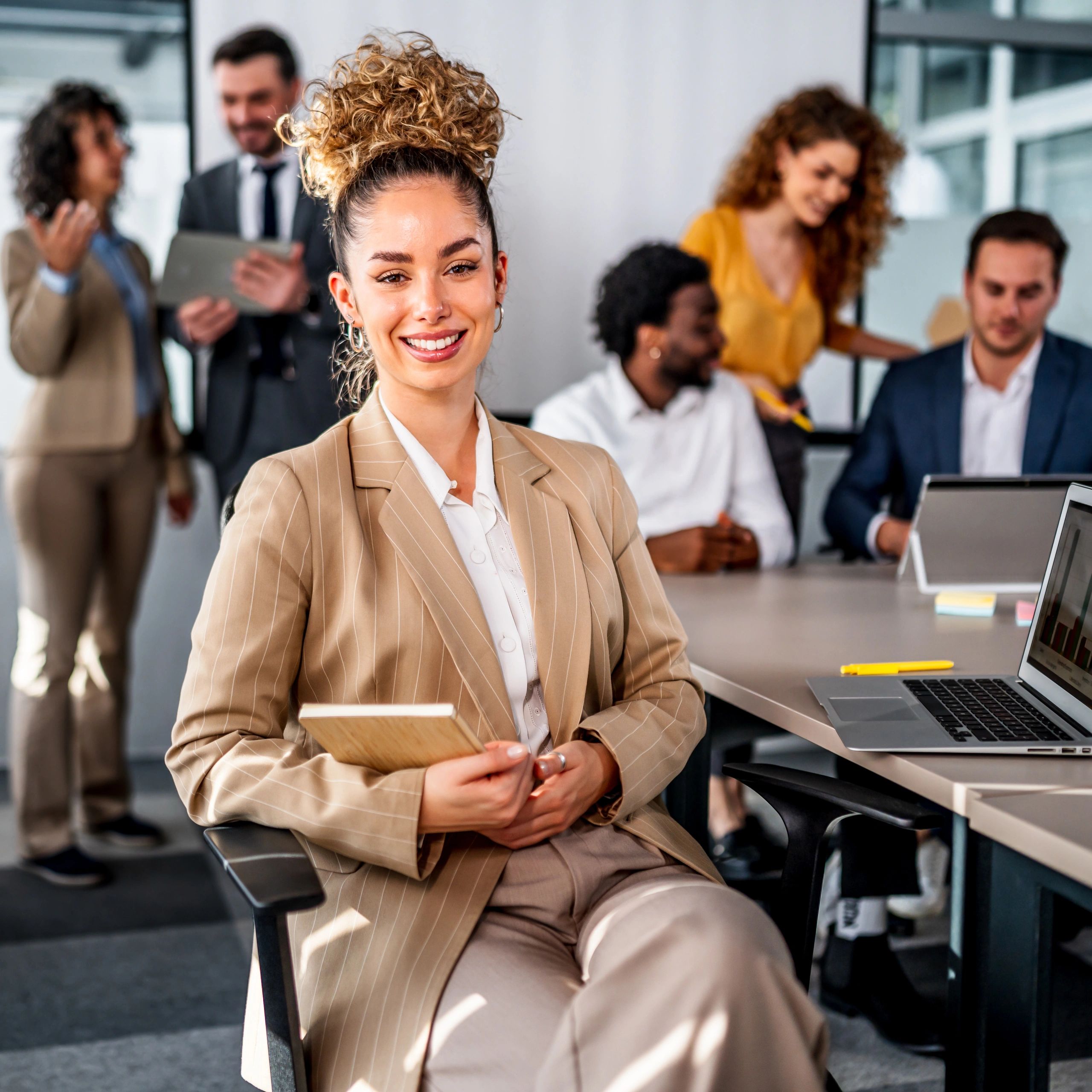 Confident businesswoman smiling in the office with colleagues collaborating in the background.