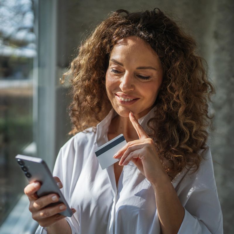 Woman standing next to a window, holding a smart phone and a credit card, smiling and looking away while contemplating online shopping or making a digital payment