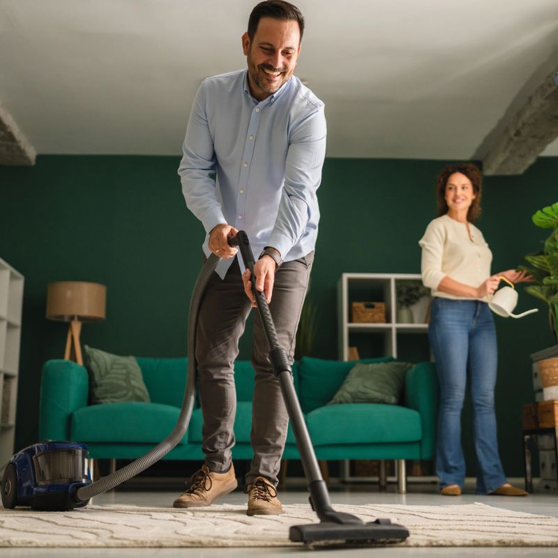 Mid adult couple engaged in domestic activities, with the man vacuuming the living room floor and the woman watering a plant, showcasing teamwork and shared responsibilities