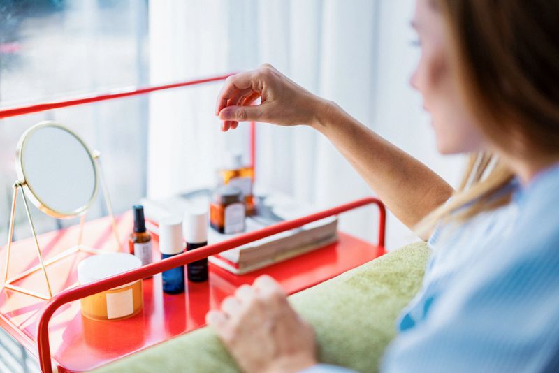 A woman reaches for a cosmetic as a small mirror and a lineup of beauty products sit on a bold red vanity tray. Natural light highlights a fresh, everyday beauty routine.