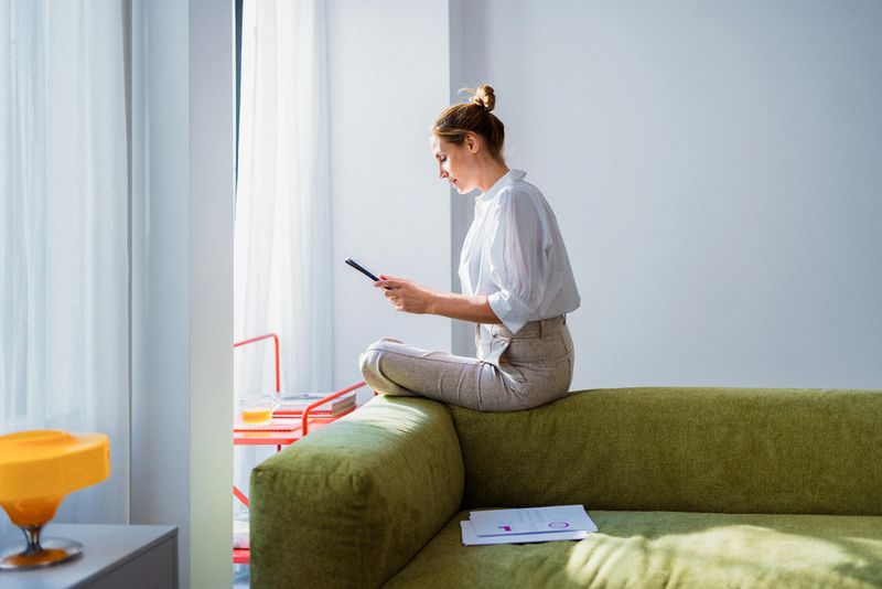 A woman sits on a green sofa in a bright living room, focused on her smartphone. Papers and a glass of juice sit nearby, suggesting a casual work-from-home moment.