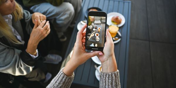 Person capturing food on a table with a smartphone while others sit nearby.