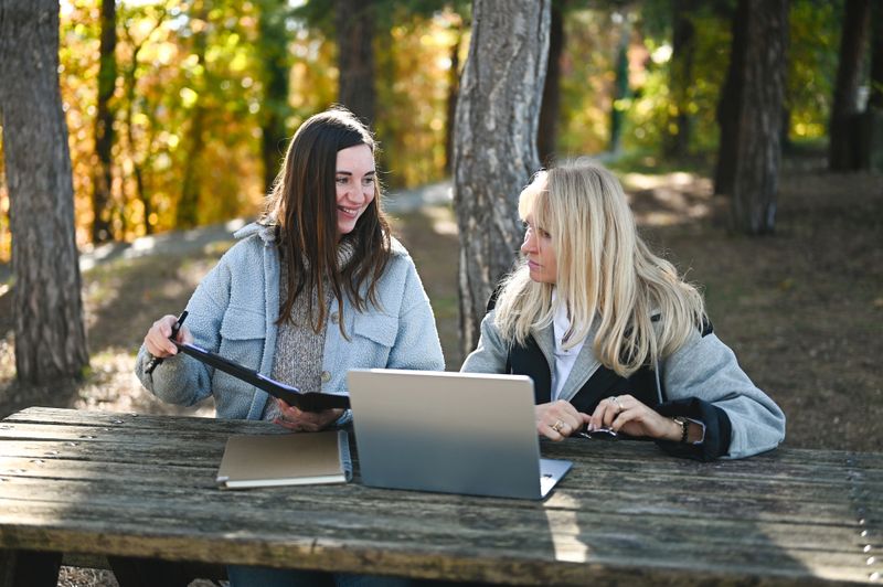 Two women discussing work over a laptop at an outdoor picnic table