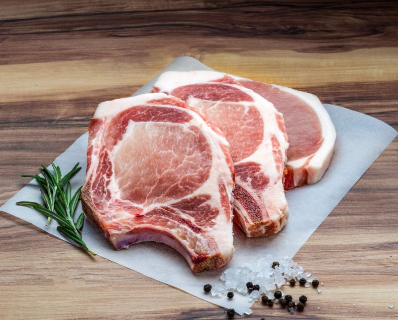 Three fresh bone-in pork chops arranged on parchment paper with rosemary sprigs, coarse sea salt, and whole black peppercorns. Ideal for culinary visuals, butcher shop promotions, recipe blogs, or food preparation content.