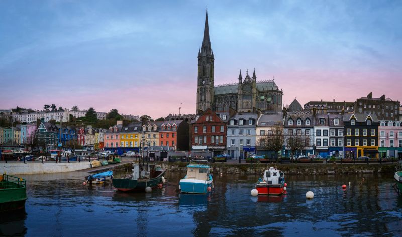 Cobh city seaport town skyline with views of St Colman's Cathedral, landmark architecture, and moored boats in Cobh Harbor, County Cork, Ireland, during an early morning sunrise as the tide rolls in.