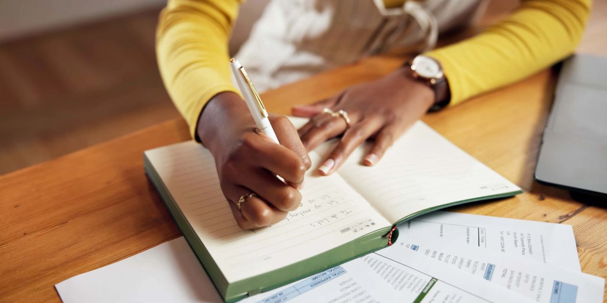 Person writing in a notebook with documents and laptop nearby.