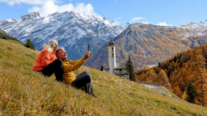 Mature couple take selfie on meadow above church and mountains in autumn, golden larch forest and snowcapped mountains distant, near Livigno and St Moritz, on the Italian-Swiss border