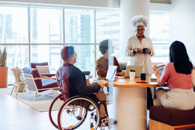 A diverse group of colleagues gathers around a round table in a modern office, discussing with laptops, mugs, and notebooks as a standing woman presents.