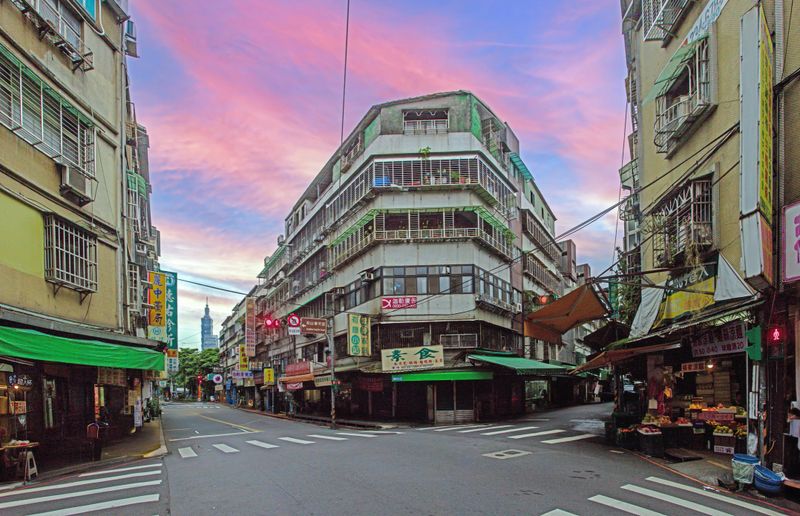 Sunset view of a quiet Taipei street lined with market stalls and residential buildings with Taipei 101 rising in the distance.