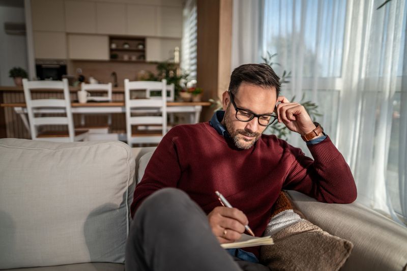 Mid-40s man wearing glasses and casual clothes, focused on journaling and planning in a cozy living room on a sofa, pen in hand, thoughtful and relaxed during the day