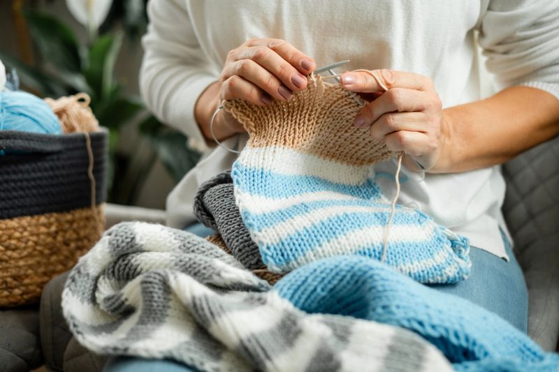 Close-up of a woman’s hands knitting a striped wool project with soft yarns in warm tones. Cozy textures, natural light, and detailed craftsmanship highlight creativity, skill, and a peaceful home atmosphere.