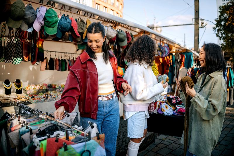 Three female friends browse and shop for clothes at a lively outdoor market. Captures friendship, casual urban lifestyle, and the vibrant atmosphere of street shopping