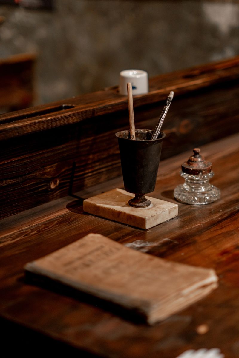 An old wooden desk features a metal holder with pens and a vintage paperweight next to a worn-out book, set in a cozy, warmly lit environment.