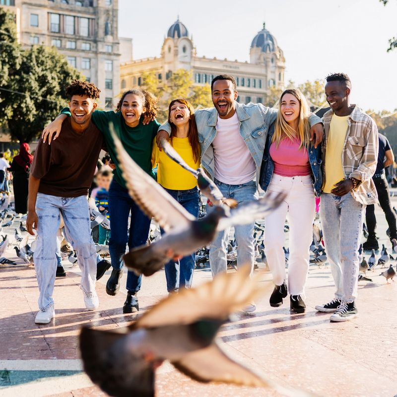 Energetic multiracial group of young millennial friends running and laughing together through Plaza Catalunya in Barcelona, Spain. Friendship and joyful lifestyle concept in an urban city atmosphere.