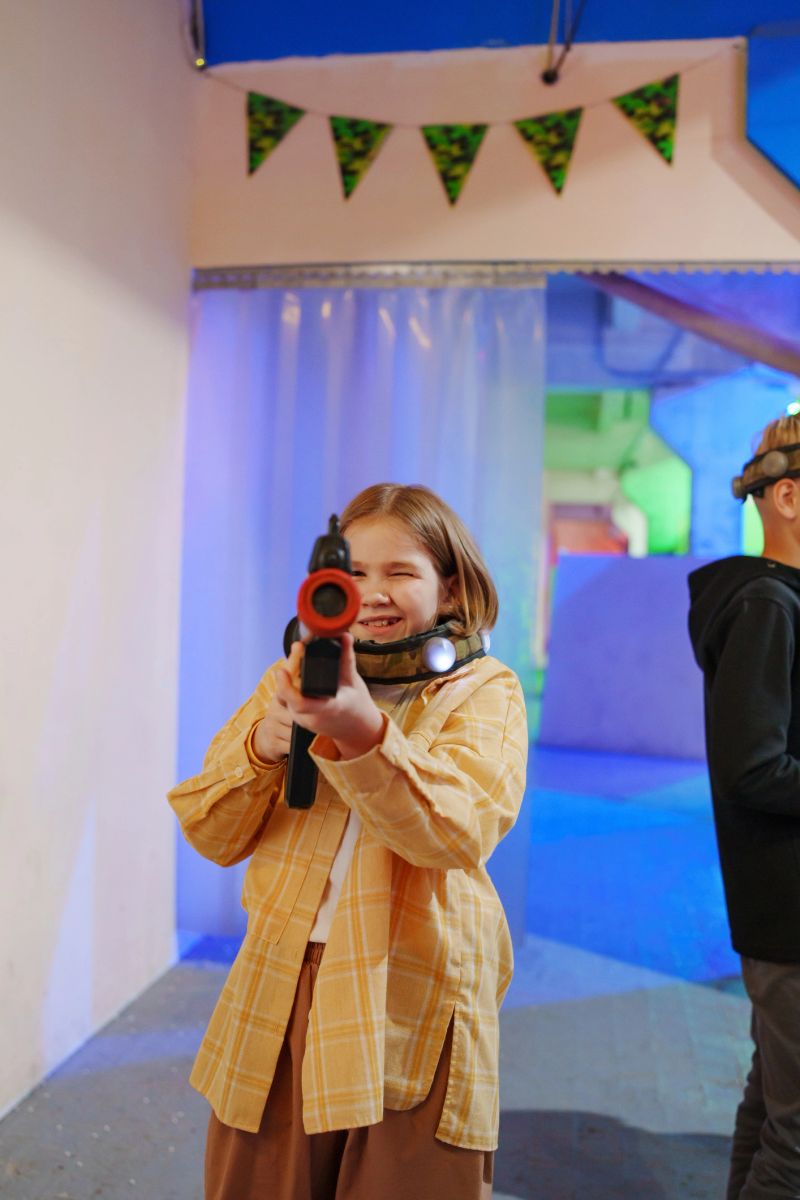A young girl holds a laser tag gun with a smile, dressed in a yellow plaid shirt. Bright colored lights and decorations fill the playful game arena.