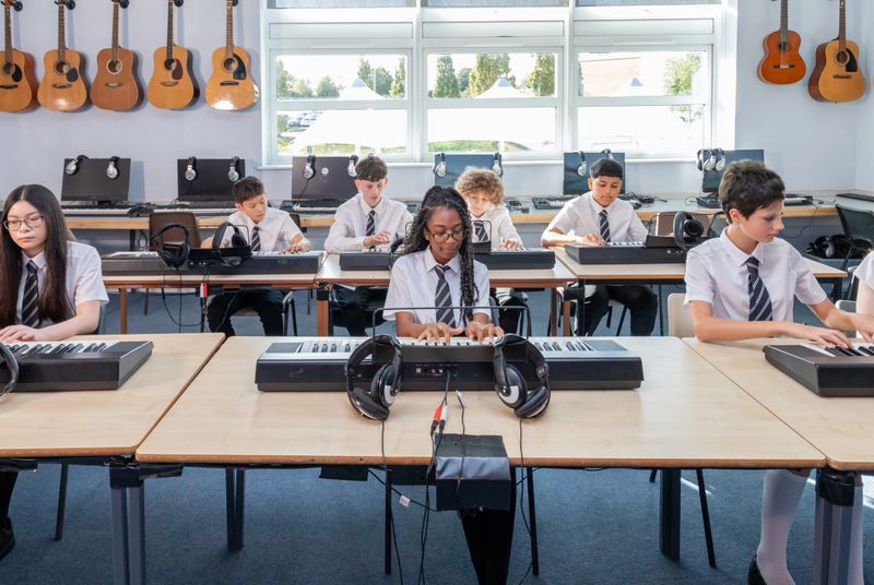 A group of students seated in two neat rows are playing electronic keyboards during a music lesson in a classroom setting. The image shows an engaged and interactive group music learning experience, with focused pupils participating in instrumental practice.